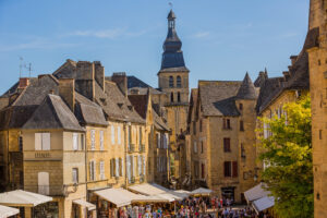 L’image montre une place vivante remplie de promeneurs et de terrasses de cafés. Les maisons en pierre dorée aux toits pentus encadrent la scène, dominée par un clocher élancé. Des auvents blancs couvrent les stands et commerces.