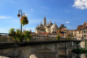 L’image montre un pont en pierre décoré de fleurs suspendues, traversant la rivière l’Isle. En arrière-plan, la cathédrale Saint-Front de Périgueux domine la ville avec ses coupoles blanches et son clocher élancé, sous un ciel bleu clair.